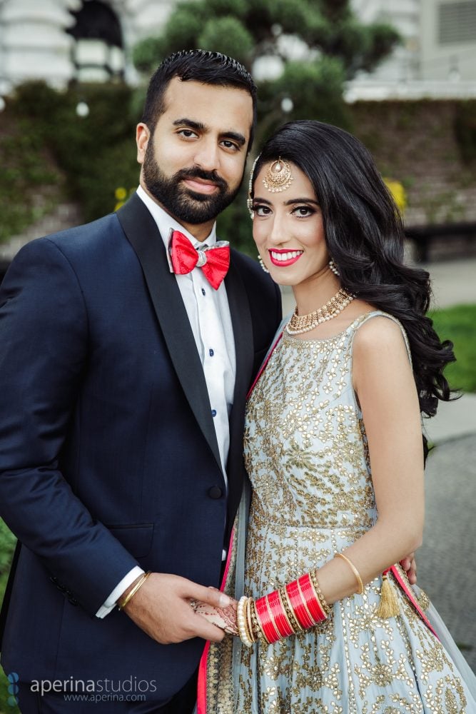Indian bride in Pratap Sons wedding dress and gold jewelry and groom with red bowtie portrait